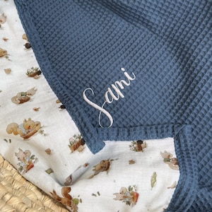 K&ouml;nnte beinhalten: Eine Decke mit zwei Texturen, bestehend aus einem dunkelblauen Waffelstrickstoff auf der einen Seite und einem wei&szlig;en Stoff mit einem wiederkehrenden Muster aus Eichh&ouml;rnchen und Herbstbl&auml;ttern auf der anderen Seite. Der Name Sami ist wei&szlig; gestickt.