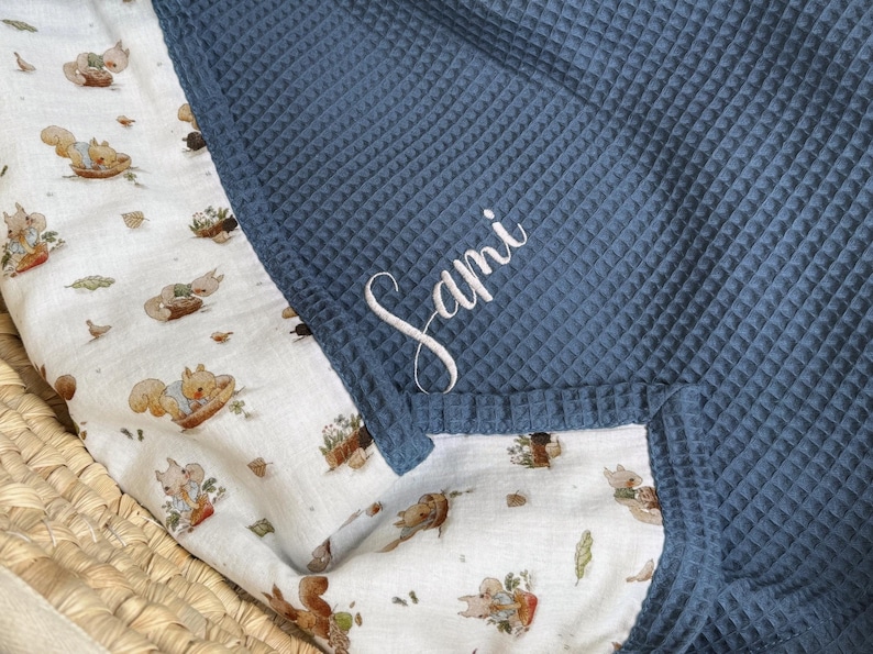 K&ouml;nnte beinhalten: Eine Decke mit zwei Texturen, bestehend aus einem dunkelblauen Waffelstrickstoff auf der einen Seite und einem wei&szlig;en Stoff mit einem wiederkehrenden Muster aus Eichh&ouml;rnchen und Herbstbl&auml;ttern auf der anderen Seite. Der Name Sami ist wei&szlig; gestickt.