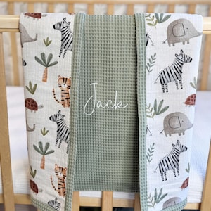 May include: A baby blanket featuring a central sage green waffle-textured panel with the name Jack embroidered in white script, flanked by two side panels of white fabric printed with a pattern of zebras, elephants, tigers, and palm trees.