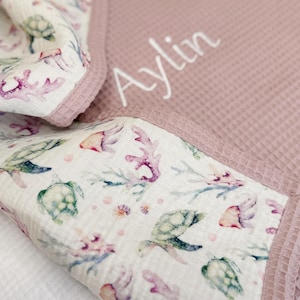 May include: A waffle-textured blanket in a dusty rose color featuring the name Aylin embroidered in white script. The blanket has a folded corner revealing a white fabric lining with a colorful watercolor print of sea turtles and coral.