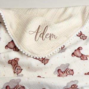 May include: A cream-colored muslin blanket featuring a repeating pattern of brown teddy bears and grey clouds with small stars. The blanket is trimmed with white pom-pom fringe and features the name Adem embroidered in brown script on the fold.