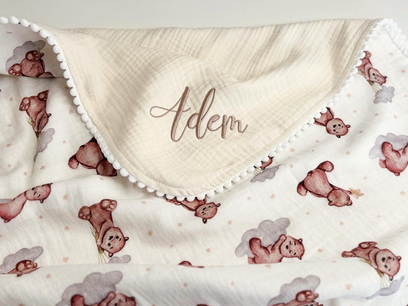 May include: A cream-colored muslin blanket featuring a repeating pattern of brown teddy bears and grey clouds with small stars. The blanket is trimmed with white pom-pom fringe and features the name Adem embroidered in brown script on the fold.