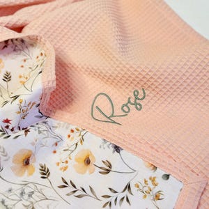 May include: A light pink waffle-textured blanket featuring a white underside with a colorful floral print of yellow, orange, and purple wildflowers. The name Rose is embroidered in dark green cursive script on the pink waffle fabric surface.