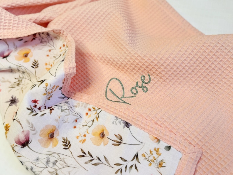 May include: A light pink waffle-textured blanket featuring a white underside with a colorful floral print of yellow, orange, and purple wildflowers. The name Rose is embroidered in dark green cursive script on the pink waffle fabric surface.