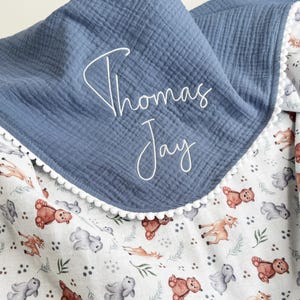May include: A blue muslin blanket featuring a white pom-pom trim and the embroidered name Thomas Jay in white script. The blanket has a white underside patterned with small illustrations of brown bears, gray rabbits, and green foliage.