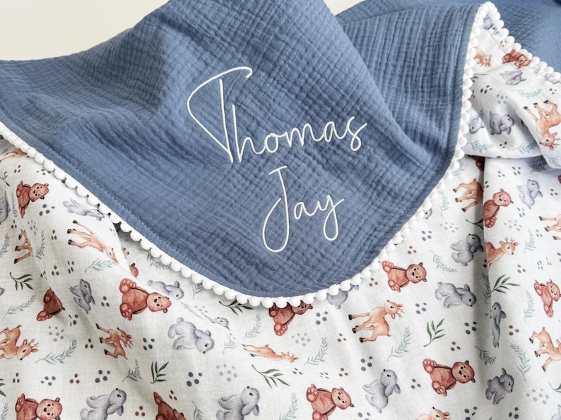May include: A blue muslin blanket featuring a white pom-pom trim and the embroidered name Thomas Jay in white script. The blanket has a white underside patterned with small illustrations of brown bears, gray rabbits, and green foliage.