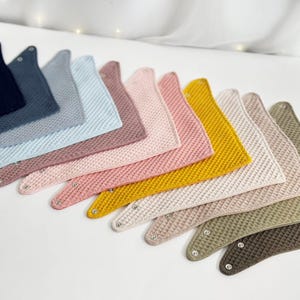 May include: A collection of waffle-textured baby bibs in various colors, including navy, blue, pink, and yellow. Each bib is triangular with snap closures at the neck, arranged in a diagonal row.