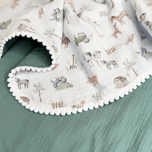 May include: A white muslin blanket featuring a repeating watercolor print of farm animals, tractors, and barns, finished with a white pom-pom trim. The blanket is draped over a solid, textured dark sage green fabric surface.