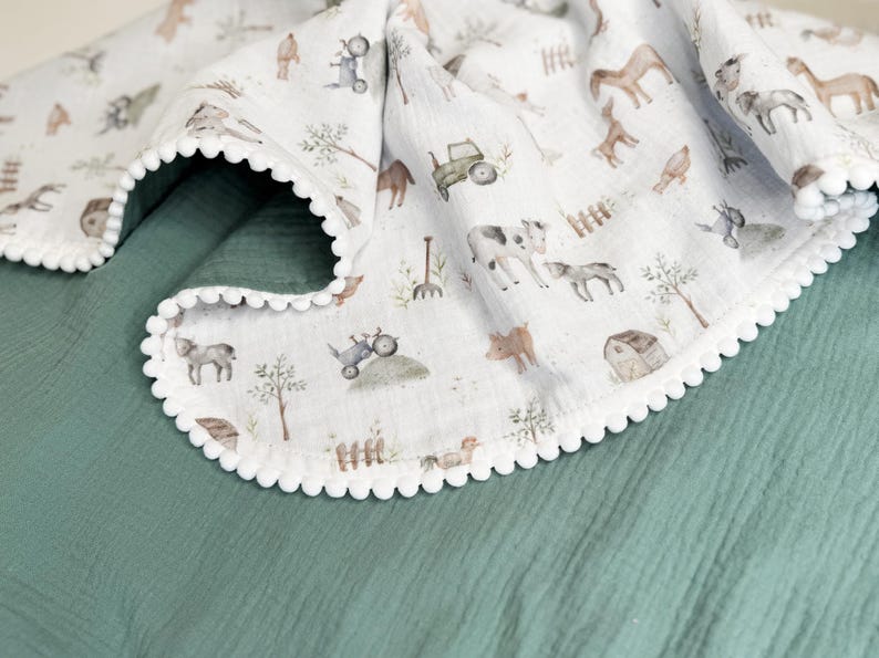 May include: A white muslin blanket featuring a repeating watercolor print of farm animals, tractors, and barns, finished with a white pom-pom trim. The blanket is draped over a solid, textured dark sage green fabric surface.