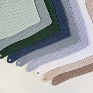 Peut inclure: Une collection de bavoirs triangulaires à texture gaufrée, disposés en éventail. Les couleurs incluent le vert sauge, le vert forêt foncé, le bleu marine, le bleu ardoise, le bleu clair, le blanc, le beige et le marron, chacun avec un bouton-pression.