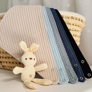 Puede incluir: Una colección de baberos para bebés de tejido de gofre en varios colores, incluyendo beige, azul claro y marino, con cierres de corchete. Un pequeño juguete de peluche con forma de conejito está delante de los baberos. Los baberos están junto a una cesta de mimbre.