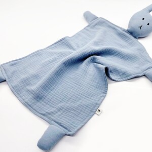 Puede incluir: Una manta de seguridad azul para beb&eacute; con una cabeza de conejo y cuatro extremidades. El conejo tiene peque&ntilde;os ojos negros y una boca cosida. La manta est&aacute; hecha de una tela arrugada. El dise&ntilde;o es simple y reconfortante.