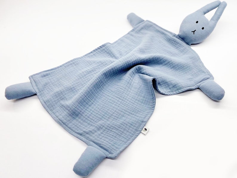 Puede incluir: Una manta de seguridad azul para beb&eacute; con una cabeza de conejo y cuatro extremidades. El conejo tiene peque&ntilde;os ojos negros y una boca cosida. La manta est&aacute; hecha de una tela arrugada. El dise&ntilde;o es simple y reconfortante.