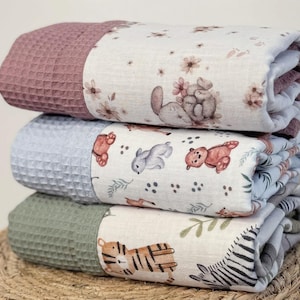 May include: Three rolled baby blankets stacked on top of each other. The top blanket is mauve with a waffle texture, the middle is light blue, and the bottom is green. Each blanket has a patterned section with animal illustrations.
