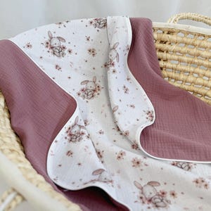 May include: A soft muslin blanket featuring a white base with a repeating pattern of small brown bunnies and pink flowers, contrasted by solid dusty rose panels on the sides. The fabric has a crinkled texture and finished white edges.