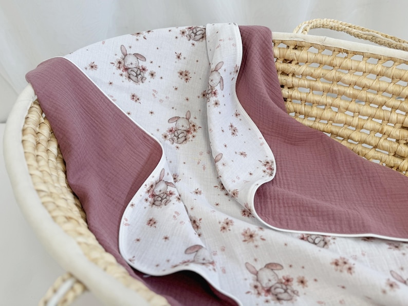 May include: A soft muslin blanket featuring a white base with a repeating pattern of small brown bunnies and pink flowers, contrasted by solid dusty rose panels on the sides. The fabric has a crinkled texture and finished white edges.