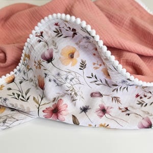 May include: A double-sided muslin blanket featuring a solid dusty rose side and a white side with a botanical print of yellow, pink, and purple wildflowers. The edges are finished with a white pom-pom trim that runs along the entire perimeter.