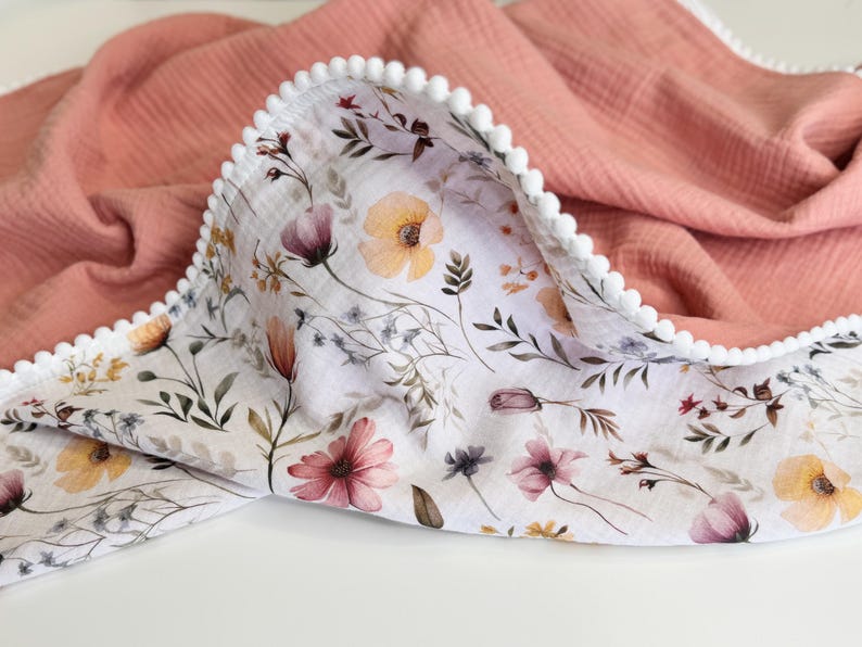 May include: A double-sided muslin blanket featuring a solid dusty rose side and a white side with a botanical print of yellow, pink, and purple wildflowers. The edges are finished with a white pom-pom trim that runs along the entire perimeter.