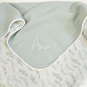 May include: A soft, light green baby blanket with a white floral pattern on one side and a solid light green on the other. The blanket has a white pom-pom trim and the name "Aria" is written in script.