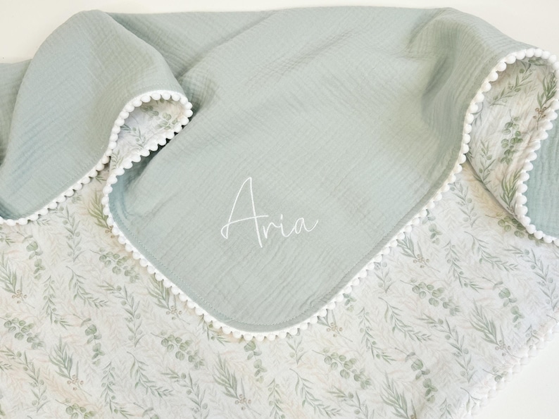May include: A soft, light green baby blanket with a white floral pattern on one side and a solid light green on the other. The blanket has a white pom-pom trim and the name "Aria" is written in script.