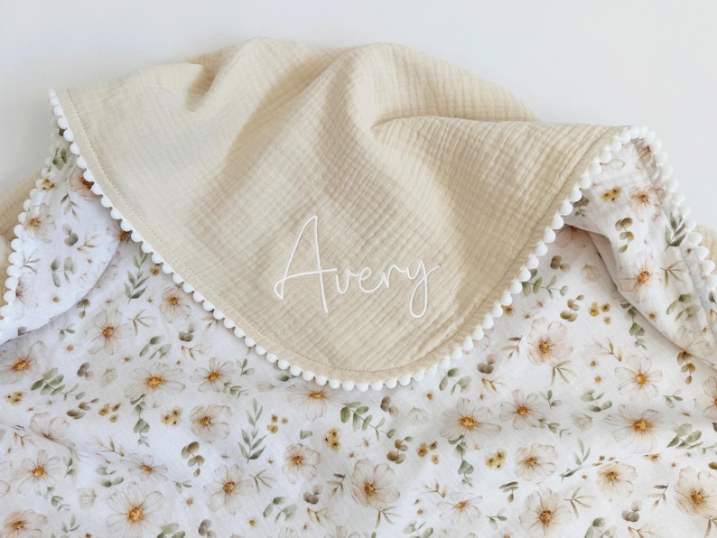 May include: A cream-colored baby blanket with a floral pattern and the name "Avery" embroidered in white. The blanket has a textured, muslin-like top layer and a white pom-pom trim.