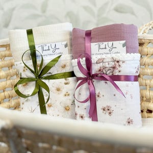 May include: Two folded muslin swaddle blankets tied with satin ribbons. One is cream with a floral print and olive ribbon, the other is mauve with a bunny and floral print and purple ribbon. Each has a tag reading Thank You for choosing Natural Cuddles.