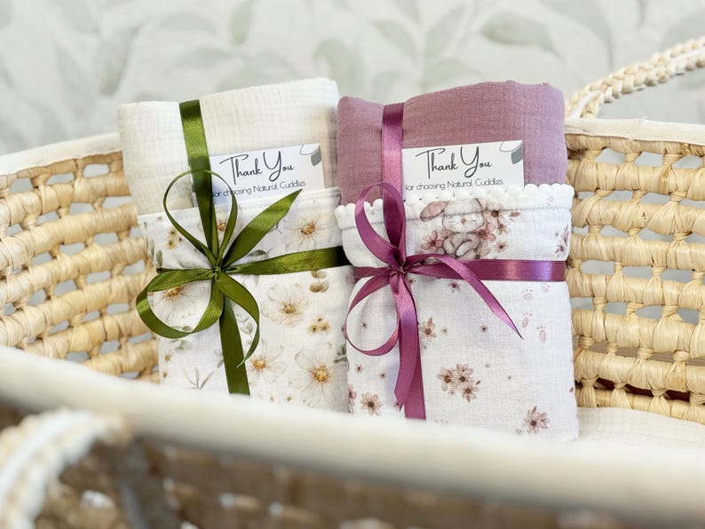 May include: Two folded muslin swaddle blankets tied with satin ribbons. One is cream with a floral print and olive ribbon, the other is mauve with a bunny and floral print and purple ribbon. Each has a tag reading Thank You for choosing Natural Cuddles.