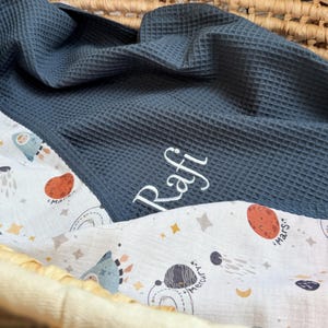 May include: A dark blue waffle-textured blanket featuring the embroidered name Rafi in white script. The blanket is lined with a white fabric patterned with colorful illustrations of planets, stars, and space-themed icons including Mars and Mercury.