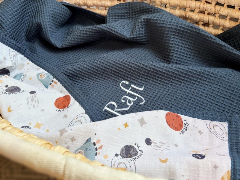 May include: A dark blue waffle-textured blanket featuring the embroidered name Rafi in white script. The blanket is lined with a white fabric patterned with colorful illustrations of planets, stars, and space-themed icons including Mars and Mercury.