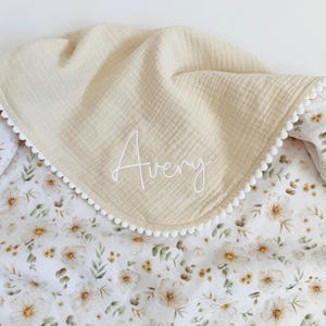 May include: A double-layered blanket featuring a solid beige muslin top side with the name Avery embroidered in white script and a white underside with a floral pattern of small brown and green flowers, finished with a white pom-pom trim.