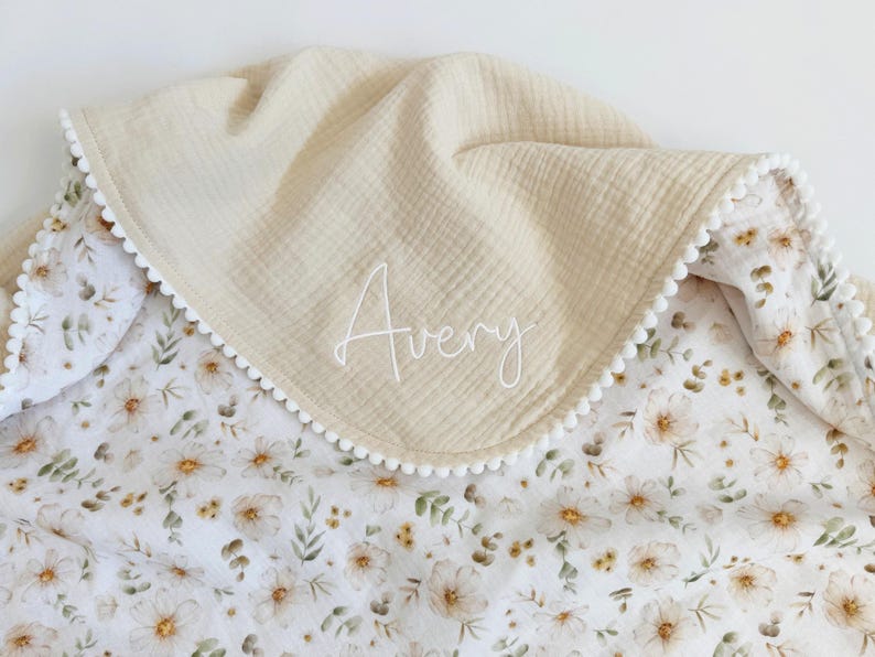 May include: A double-layered blanket featuring a solid beige muslin top side with the name Avery embroidered in white script and a white underside with a floral pattern of small brown and green flowers, finished with a white pom-pom trim.