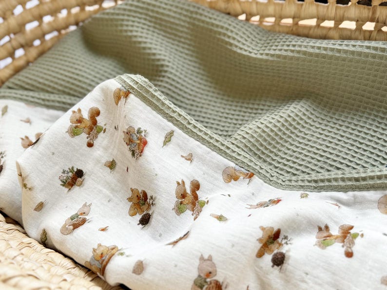 May include: A dual-textured blanket featuring a white fabric side with a repeating pattern of brown squirrels, acorns, and small green leaves, paired with a solid sage green waffle-knit fabric side, folded to display both distinct material surfaces.