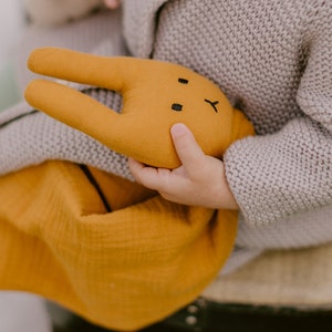 Puede incluir: Un juguete de peluche de conejo amarillo mostaza con detalles faciales negros es sostenido por un ni&ntilde;o. El conejo se combina con una manta de muselina a juego. El ni&ntilde;o lleva un su&eacute;ter de punto gris.
