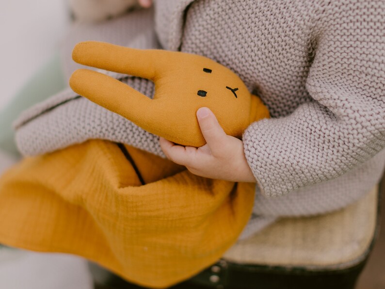 Puede incluir: Un juguete de peluche de conejo amarillo mostaza con detalles faciales negros es sostenido por un ni&ntilde;o. El conejo se combina con una manta de muselina a juego. El ni&ntilde;o lleva un su&eacute;ter de punto gris.