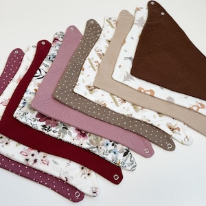 May include: A collection of baby bibs in various colors and patterns. Designs include polka dots, floral prints, and woodland scenes with foxes and trees. Bibs are made of soft fabric and have snap closures.