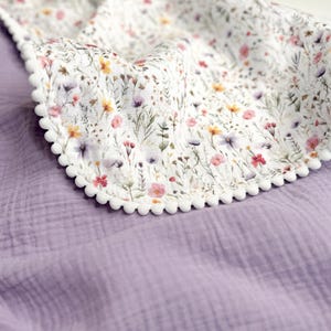 May include: A double-layered blanket featuring a solid lavender muslin fabric on one side and a white fabric with a colorful wildflower print on the other. The edges of the floral section are finished with a decorative white pom-pom trim.