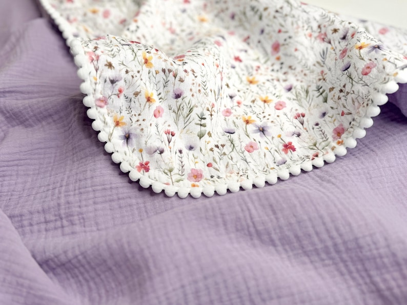 May include: A double-layered blanket featuring a solid lavender muslin fabric on one side and a white fabric with a colorful wildflower print on the other. The edges of the floral section are finished with a decorative white pom-pom trim.