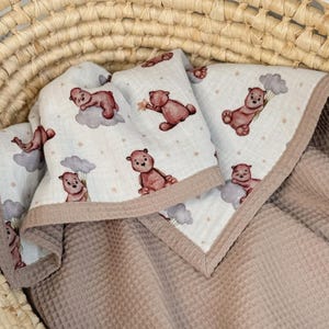 K&ouml;nnte beinhalten: Eine weiche, mehrlagige Decke mit einer wei&szlig;en Oberseite, die mit einem wiederkehrenden Muster aus braunen Teddyb&auml;ren, grauen Wolken und kleinen Sternen bedruckt ist. Die Unterseite und der Rand bestehen aus strukturiertem, hellbraunem Waffelstoff.
