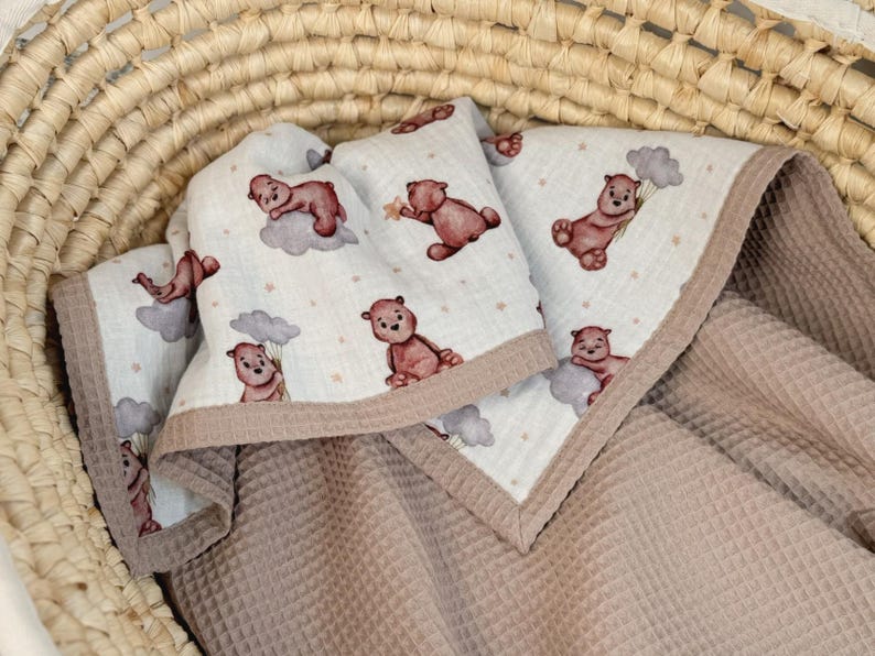 K&ouml;nnte beinhalten: Eine weiche, mehrlagige Decke mit einer wei&szlig;en Oberseite, die mit einem wiederkehrenden Muster aus braunen Teddyb&auml;ren, grauen Wolken und kleinen Sternen bedruckt ist. Die Unterseite und der Rand bestehen aus strukturiertem, hellbraunem Waffelstoff.