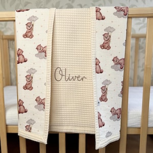 May include: A cream-colored waffle-knit baby blanket featuring a central panel embroidered with the name Oliver in brown script. The side panels display a repeating pattern of brown bears holding gray clouds against a cream background with dots.