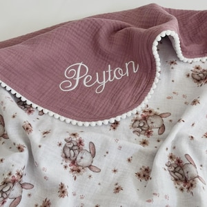 May include: A dusty rose baby blanket with a white pom-pom trim and the name "Peyton" embroidered in white script. The reverse side features a white floral pattern with bunnies and flowers.