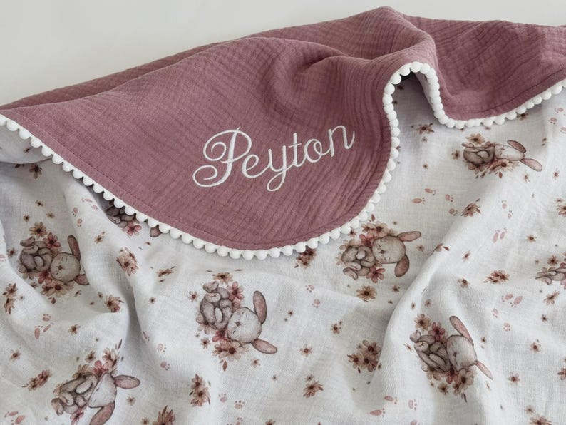 May include: A dusty rose baby blanket with a white pom-pom trim and the name "Peyton" embroidered in white script. The reverse side features a white floral pattern with bunnies and flowers.