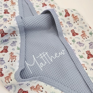 May include: A baby blanket with a blue waffle-weave hood and trim. The blanket features a repeating pattern of deer, bears, and rabbits in shades of brown, red, and grey on a white background. The name "Matthew" is written on the hood.