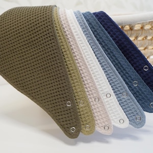May include: A collection of seven triangular bibs featuring a waffle-knit texture, arranged in a gradient of colors including olive green, sage, cream, white, light blue, slate blue, and navy blue, each secured with silver-toned metal snap buttons.