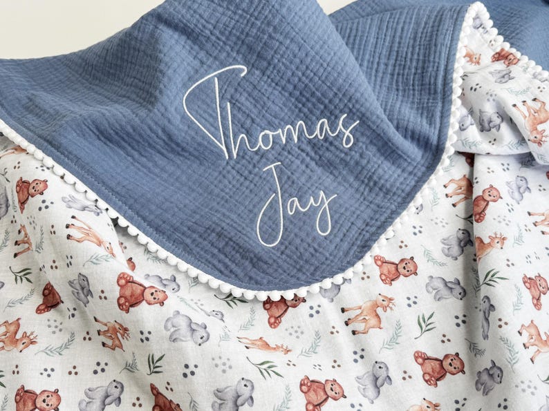 May include: A blue and white baby blanket with the name "Thomas Jay" embroidered in white script. The blanket features a blue top layer and a white layer with a pattern of bears, deer, and other forest animals. The edge is trimmed with white pom-poms.