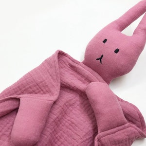 Puede incluir: Un arrullo con forma de conejito rosa para beb&eacute;s. El peluche tiene orejas largas, una cara cosida y un cuerpo de manta arrugada. El arrullo est&aacute; hecho de una tela suave y texturizada.
