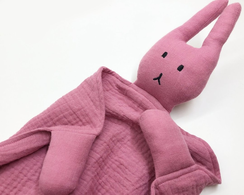 Puede incluir: Un arrullo con forma de conejito rosa para beb&eacute;s. El peluche tiene orejas largas, una cara cosida y un cuerpo de manta arrugada. El arrullo est&aacute; hecho de una tela suave y texturizada.