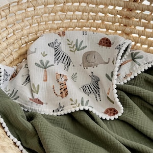 May include: A baby blanket with a white border and a green backing. The top layer features a safari animal print with zebras, elephants, and tigers. The blanket is nestled in a woven basket.