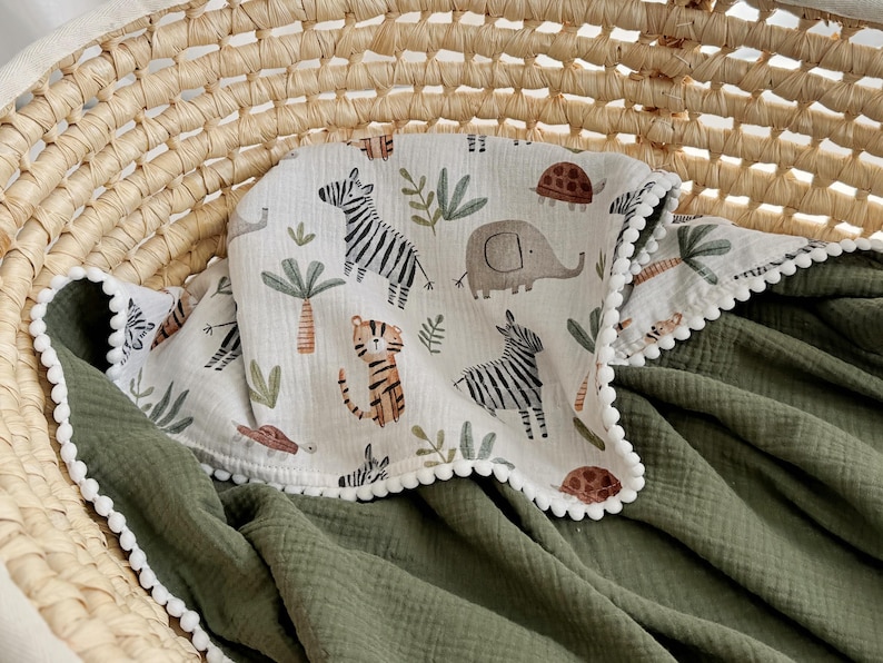 May include: A baby blanket with a white border and a green backing. The top layer features a safari animal print with zebras, elephants, and tigers. The blanket is nestled in a woven basket.