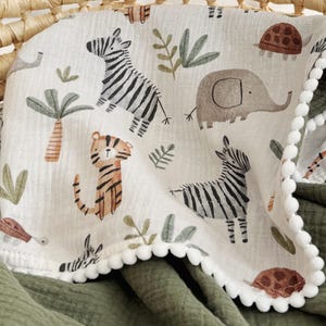 May include: A cream-colored muslin blanket featuring a repeating pattern of zebras, elephants, tigers, turtles, and palm trees. The blanket is trimmed with small white pom-poms along the edge and is layered over a solid olive green fabric.